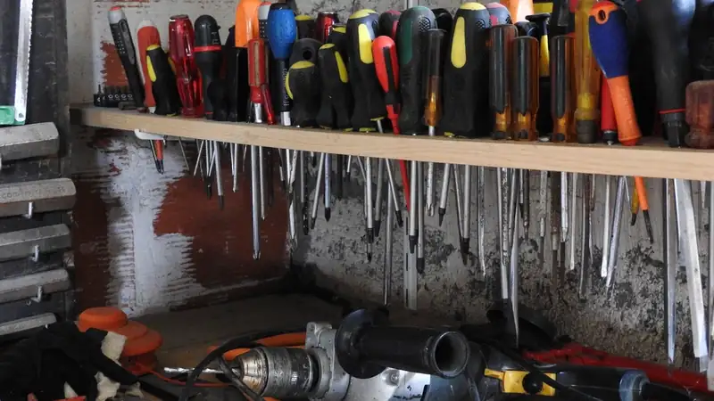 A shelf packed with screwdrivers above a cluttered workbench holding a corded drill and cables.