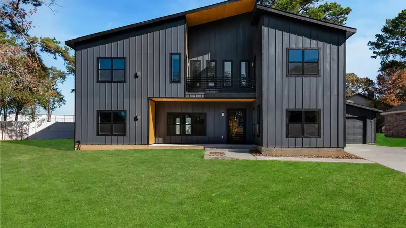 A two-story house with dark vertical siding, wood accents and a wide green front lawn.