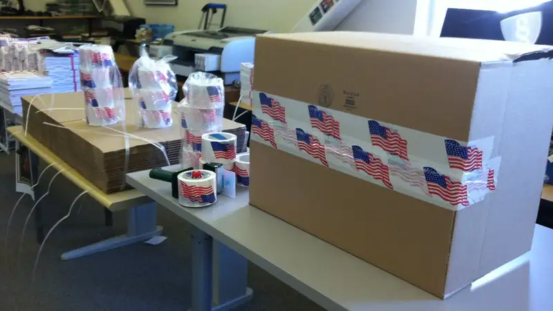 A cardboard shipping box sealed with flag-print tape on a packing table beside rolls of tape.