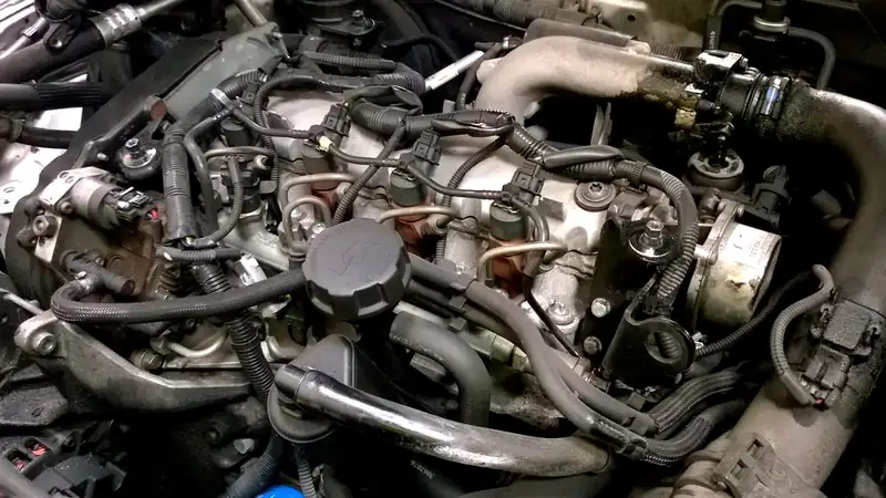 A car engine bay seen from above, crowded with hoses, wiring and the oil filler cap.