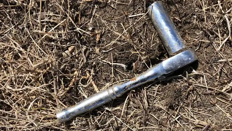 A chrome ratchet wrench with a socket attached, lying in dry grass and dirt.