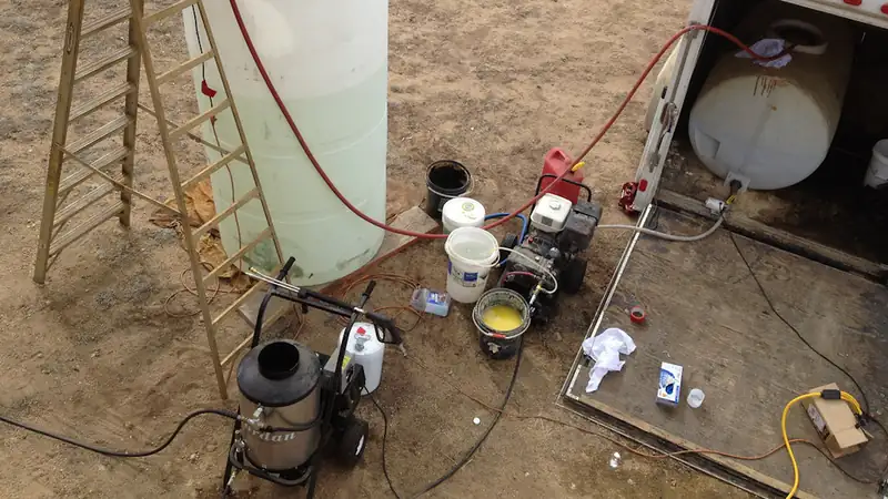 A pressure washer, hoses, buckets and a water tank set up on bare ground beside an open utility trailer.