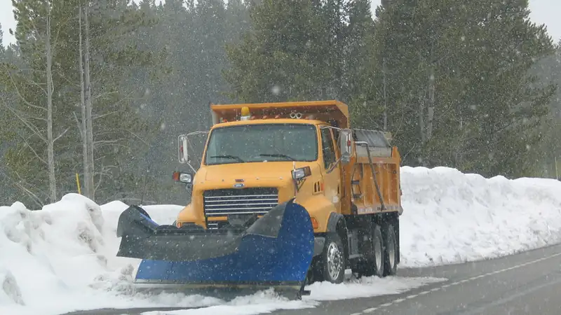 An orange dump truck with a blue plow blade parked beside high snowbanks on a forest road.