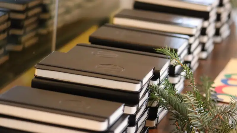 Stacks of black notebooks lined up on a table beside a sprig of evergreen.
