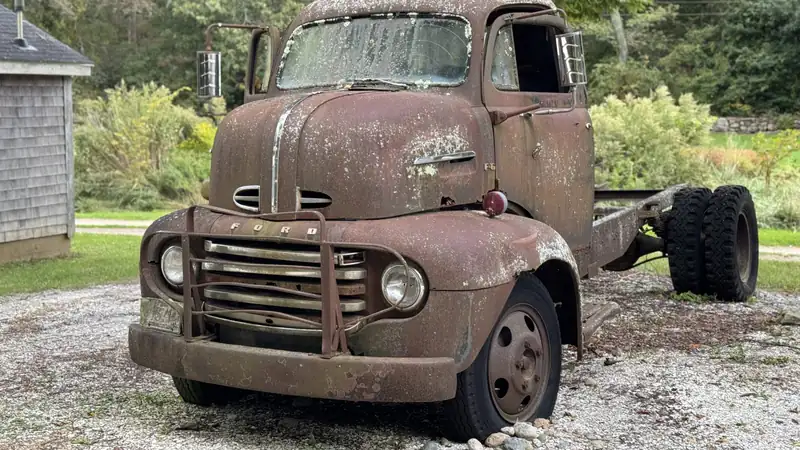 A weathered vintage cab-over truck with a bare flatbed chassis parked on a gravel driveway.