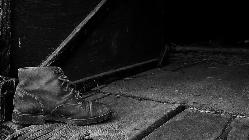 A worn leather work boot on a weathered wooden floor in black and white.